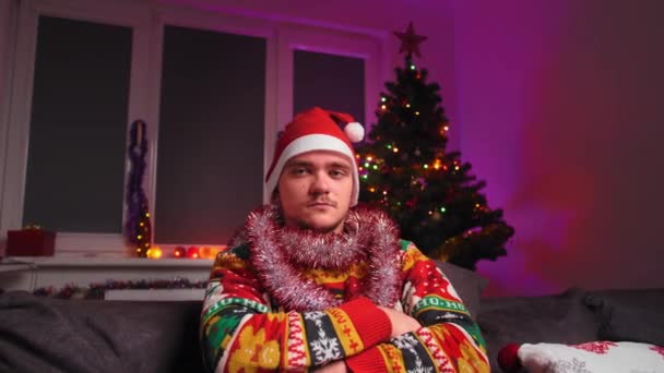 남자가 소파에 앉아서 카메라를 있습니다 크리스마스 스웨터를 모자를 나무가 있습니다 — 비디오