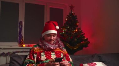 Genç bir adam kanepede oturuyor ve bir el çırpma tahtası çırpıyor, bir Noel süveteri ve şapka giyiyor, yanında bir yılbaşı ağacı, bir noel ağacı ve yeni yıl konsepti var.