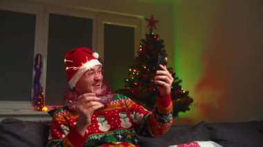 Genç bir adam video bağlantısıyla konuşuyor ve bir maytap tutuyor, bir Noel süveteri ve şapka giyiyor, yanında bir yılbaşı ağacı, bir noel ağacı ve yeni yıl konsepti var.