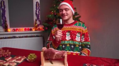 Genç bir adam festival masasına oturur ve kameraya bakar, elinde bir bardak şarap, üzerinde Noel süveteri ve şapkası, yanında bir yılbaşı ağacı, bir Noel konsepti vardır.