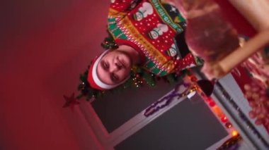 Genç bir adam festival masasına oturur ve kameraya bakar, elinde bir bardak şarap, üzerinde Noel süveteri ve şapkası, yanında bir yılbaşı ağacı, bir Noel konsepti vardır.