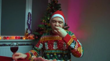 Genç bir adam şenlik masasında oturmuş jamon yiyor ve şarap içiyor, Noel süveteri ve şapka giyiyor, yanında yeni yıl ağacı, Noel ve yeni yıl konsepti var.