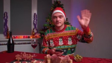 Genç bir adam festival masasında oturuyor ve kameraya bakıyor elinde bir telefon ve kameraya el sallıyor, bir Noel kazağı ve şapka giyiyor, yanında bir yılbaşı ağacı var.