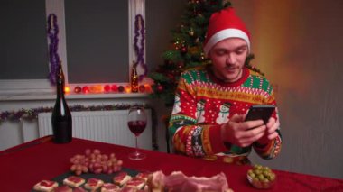 Genç bir adam festival masasında oturuyor ve telefonuyla video iletişimiyle konuşuyor, Noel süveteri ve şapkası giyiyor, yanında bir yılbaşı ağacı, bir Noel ve yeni yıl konsepti var.
