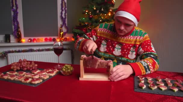 若い男はお祝いのテーブルに座って ジャモンをカットし 彼はクリスマスセーターと帽子を着ています 彼の隣には新年の木 クリスマスと新年の概念です — ストック動画