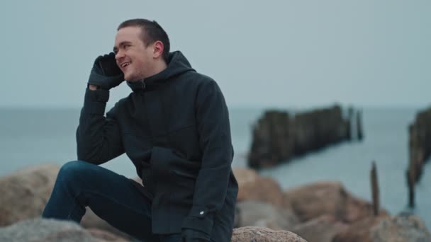 一个年轻人正靠着蓝色的大海坐在一块石头上 — 图库视频影像