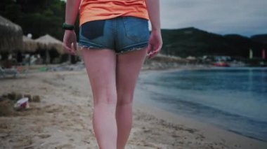 Şemsiyelerin ve şemsiyelerin yattığı deniz kıyısında yürüyen kadın, kamera ayaklarından kapanışa doğru hareket eder, akşam. Yüksek kalite 4k görüntü