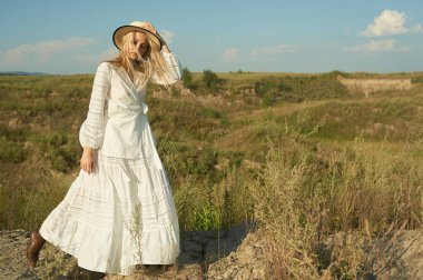 Beyaz romantik elbiseli, yaz mevsiminde muhteşem bir genç kadın. Sarı çimenler, mavi gökyüzü. Güneşli bir gün. Büyük güzel gözleri ve güzel dudakları olan sarışın bir manken. Sakin ve dengeli. Şık görünüş.  