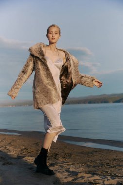 Deniz tabanında kumsalda yürüyen muhteşem sarışın kadın. Güzel, uzun boylu, ince vücutlu. İpek gri elbise, yapay kürk manto. Siyah çizmeler. Gün batımı. Mavi gökyüzü. Romantik ruh hali.  