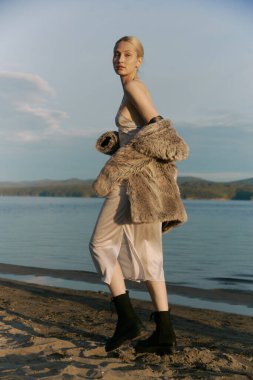 Deniz tabanında kumsalda yürüyen muhteşem sarışın kadın. Güzel, uzun boylu, ince vücutlu. İpek gri elbise, yapay kürk manto. Siyah çizmeler. Gün batımı. Mavi gökyüzü. Romantik ruh hali.  