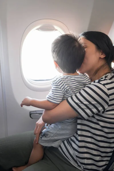 Infant traveling in airplane sitting on its mother lap both looking out of an airplane window. One year old baby boy flying in airplane and trying to entertain himself