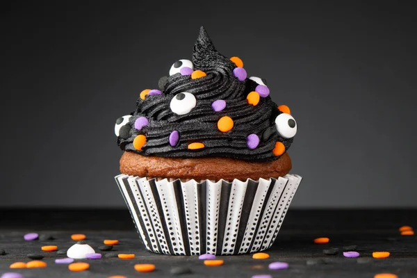 Cupcake on Halloween. Dessert on Halloween party. Muffin decorated with colored sprinkles, black frosting, icing. Cupcake on dark background and wooden desk. Macro high quality and resolution photo.