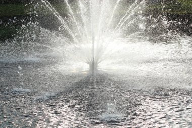On a hot day, there is a beautiful fountain in the park. Splashes of water from the fountain in the backlight.