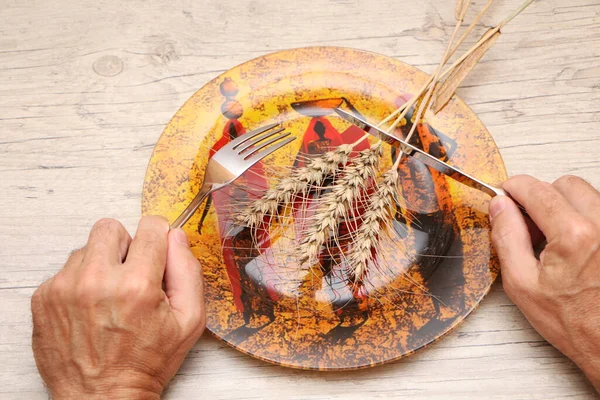 On a plate with African patterns is a grain ear, in the hands of a fork and a knife. A symbol of hunger and inept housekeeping is a plate with cutlery and a spikelet against the background of a wooden structure.