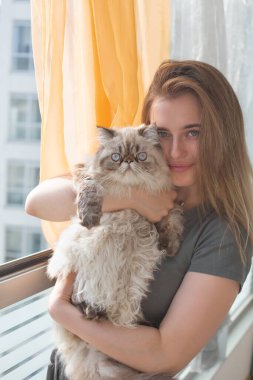 Genç ve güzel bir kız, Pers kedisini kollarında tutuyor ve pencerenin yanında dururken gülümsüyor. Kollarında bir kız ve bir kedi kameranın lensine bakıyor..