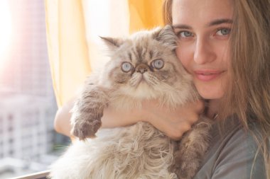 Genç ve güzel bir kız, Pers kedisini kollarında tutuyor ve pencerenin yanında dururken gülümsüyor. Kollarında bir kız ve bir kedi kameranın lensine bakıyor..