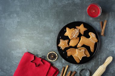Siyah bir tabakta çeşitli şekillerde Noel kurabiyeleri, mutfak kırmızı havlusu ve diğer mutfak malzemeleri koyu gri arka planda bulunmaktadır. En üst noktadan bak. Metin için boş alan.
