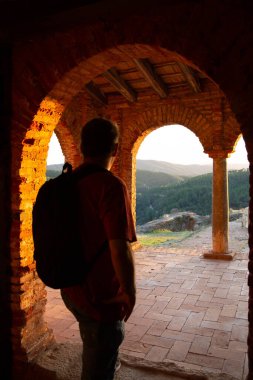 Sırt çantalı bir erkek silueti gün batımında aydınlatılmış eski bir kırmızı tuğla binanın kemerinin önünde duruyor. Seyahat teması.