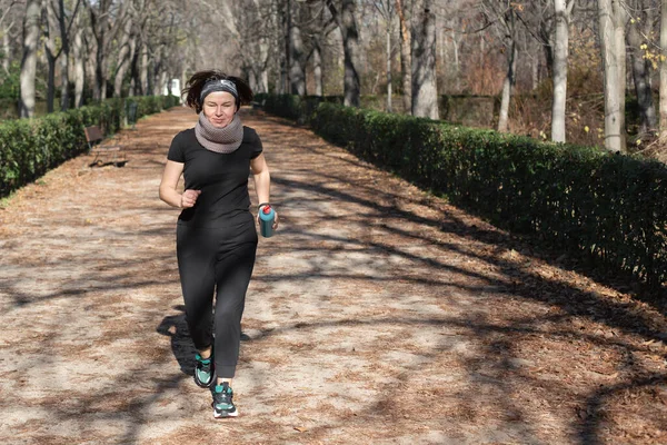 Siyah spor giysili bir kadın parkın sonbaharında yolun aşağısında koşuyor. Sağlıklı bir yaşam tarzı ve spor teması.