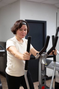 Orta yaşlı esmer bir kadın, spor salonunda eliptik antrenör kullanıyor. Bir kadın simülatördeki bacak kaslarını güçlendirir..