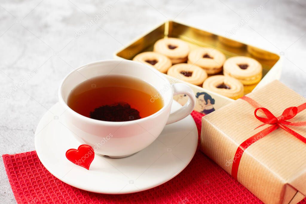 A white cup of tea, a gift packed with red ribbon and a box of cookies are on the table. Theme breakfast on Valentine's Day.