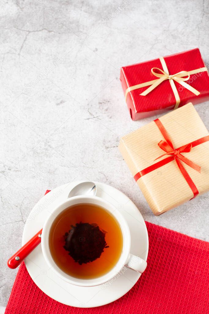 A white cup of tea with spoon and two packaged gifts with red ribbon are on gray background. Theme breakfast on Valentine's Day. View from the top point. A vertical image.