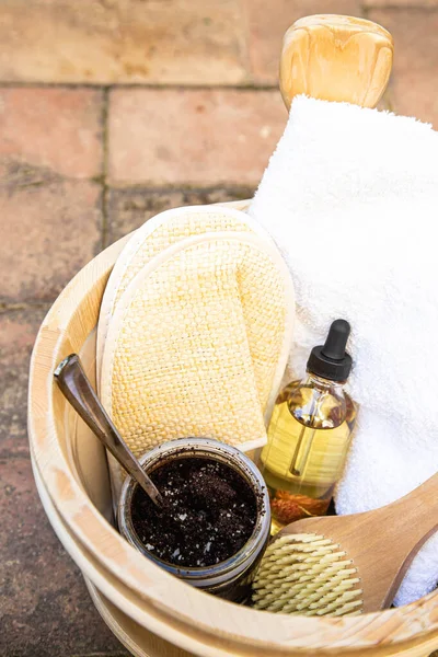 A coffee scrub with salt, glass jar with organic oil, wooden brush and white bath towel are on a wooden bucket. The theme of natural cosmetics and sauna. Top view or flat lay. A vertical image.