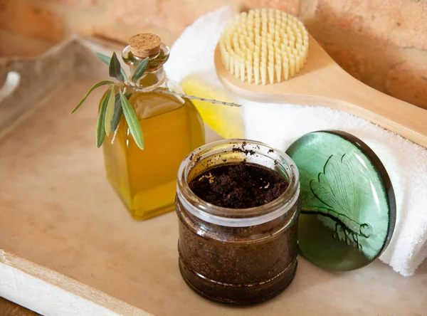 A jar of handmade coffee scrub, body brush with bottle of olive oil and white towel are on wooden tray. Free space for text.