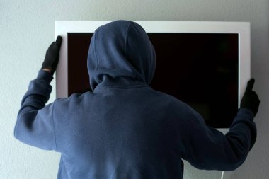 Kapüşonlu sweatshirt ve siyah eldivenli bir adam televizyonunu duvardan indiriyor. Hırsızlık ve kriminoloji teması.