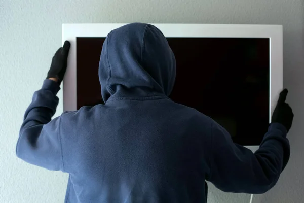 Kapüşonlu sweatshirt ve siyah eldivenli bir adam televizyonunu duvardan indiriyor. Hırsızlık ve kriminoloji teması.