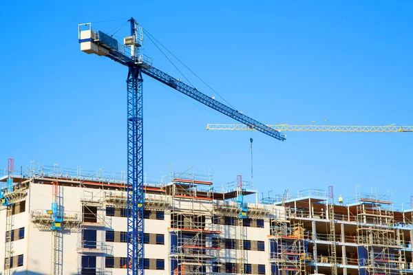 A building under construction with builders on the roof and cranes stand against a blue sky.