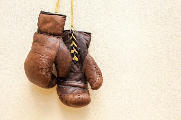 Old worn leather brown boxing gloves hang against the background of the wall in yellow. Free space for text.