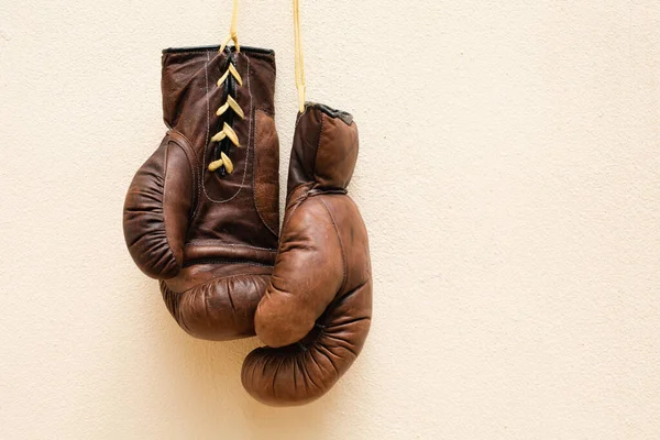 Old worn leather brown boxing gloves hang against the background of the wall in yellow. Free space for text.