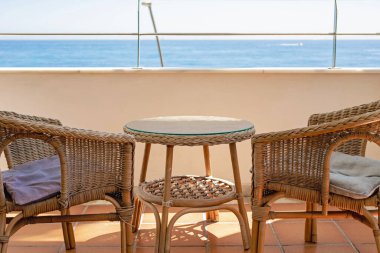 Two wicker chairs and a round table are located on the terrace against the background of the blue sea.