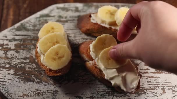 Προετοιμασία πρωινό φρούτα, γυναίκα χέρι βάζει μπανάνα φέτα σε σάντουιτς με τυρί κρέμα, κοντά — Αρχείο Βίντεο