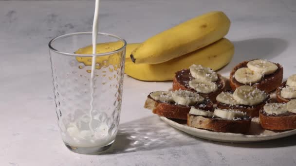 Солодкий сніданок, заливання молока в склянку, бананові бутерброди з шоколадом на фоні — стокове відео