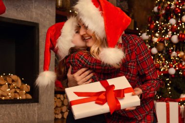 Güzel, mutlu anne ve Noel Baba şapkaları ve Noel Baba kırmızı gömlekleri içinde şirin bir kız birbirlerine hediye kutuları, şömine ve Noel ağacı arka planında bir hediye kutusu veriyorlar.