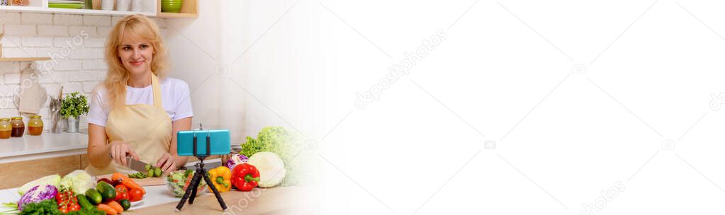 A beautiful happy young girl is blogging for her kitchen channel about a healthy lifestyle and new recipes in the kitchen of her home and looking at the smartphone camera on a tripod with a smile