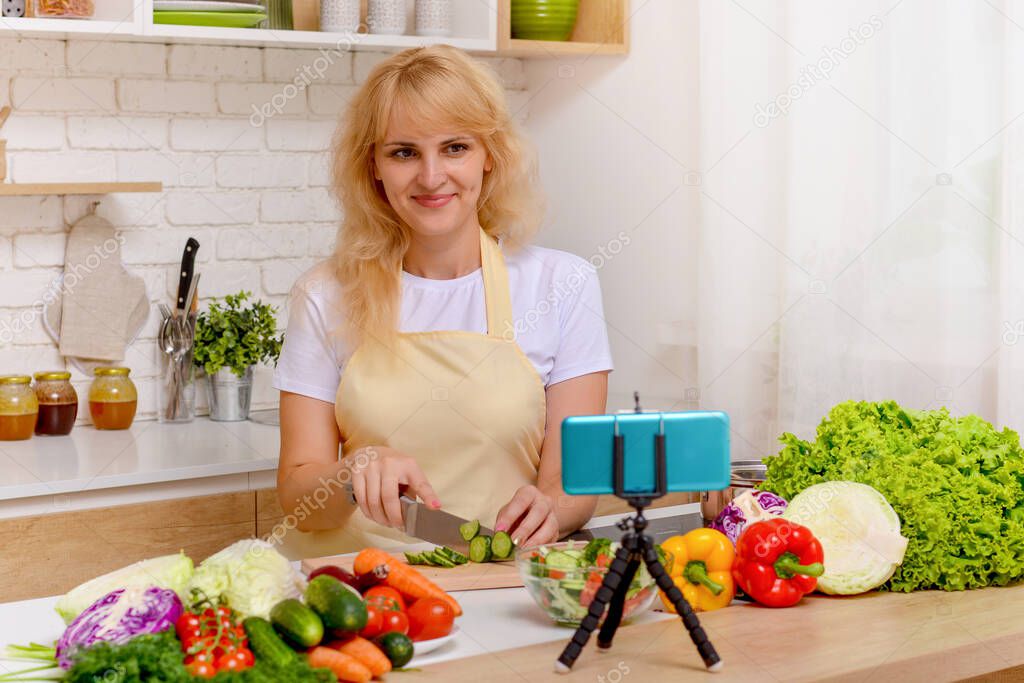 A beautiful happy young girl is blogging for her kitchen channel about a healthy lifestyle and new recipes in the kitchen of her home and looking at the smartphone camera on a tripod with a smile