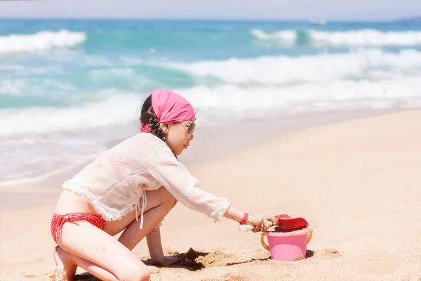 A beautiful girl plays with sand on the beach on the backdrop of the sea or ocean during a beach holiday. The kid builds sand paska with a bucket and spatula. Children\'s Games on the beach.