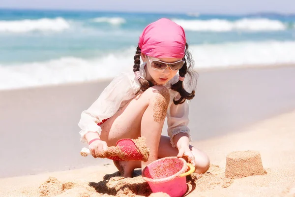A beautiful girl plays with sand on the beach on the backdrop of the sea or ocean during a beach holiday. The kid builds sand paska with a bucket and spatula. Children's Games on the beach.
