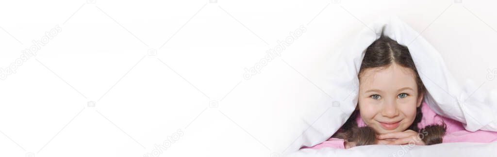 Portrait of a happy cute girl lying on white bed linen under a blanket. Banner. Copy space for text
