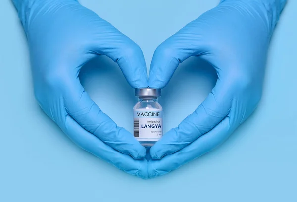 Doctor\'s hands in protective medical gloves hold a bottle of Langya henipavirus (LayV) vaccine in a heart-shaped gesture on a blue background. Vaccination. Immunization. Fighting the pandemic.
