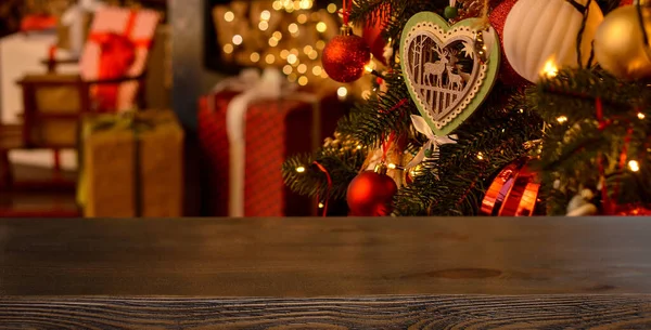 Empty wooden table on the background of a Christmas tree with beautiful toys in the form of balls, a wooden heart with deer, garlands and boxes with gifts, fireplace with golden bokeh..Mockup