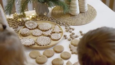 Anne ve çocuklar mutfakta Noel kurabiyesi süslerler. Bir erkek ve bir kız, kurabiyelerin üzerine şeker kremalı korselerle resim yapıyorlar. Yılbaşı şekerleri Noel ağacının dalları.
