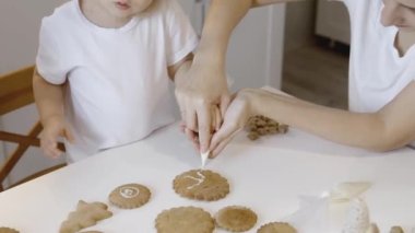 Anne ve çocuklar Noel kurabiyelerini evde süslerler. Bir erkek ve bir kız, kurabiyelerin üzerine şeker kremalı korselerle resim yapıyorlar. Noel ağacının yeni yıl süslemeleri..