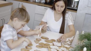 Anne ve çocuklar Noel kurabiyelerini evde süslerler. Bir erkek ve bir kız, kurabiyelerin üzerine şeker kremalı korselerle resim yapıyorlar. Noel ağacının yeni yıl süslemeleri..