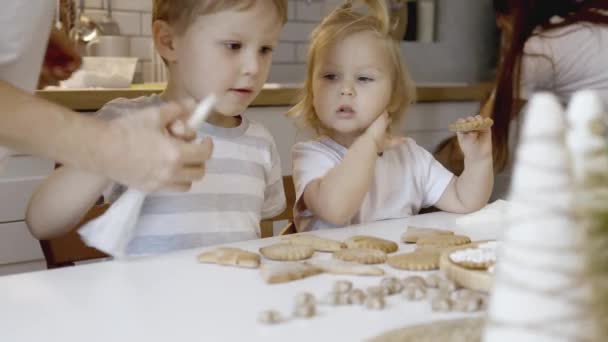 ママと子供たちは自宅でクリスマスジンジャーブレッドを飾る 男の子と女の子がクッキーに砂糖アイシングでコーネットでペイントします クリスマスツリーの新年の台所の装飾枝 — ストック動画