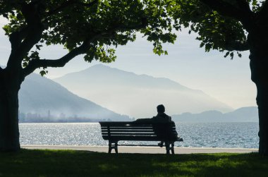 Silüet içinde sırtüstü oturmuş, sabah sakin bir göle bakıyor. Arka planda dağlar ve sis var..
