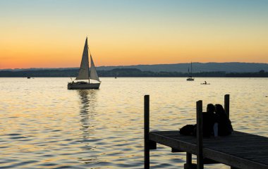 Gün batımında Zugersee Gölü 'nde manzara. İsviçre' nin Zug kasabasında, ufuktan geçen bir yelkenli, gün batımını izleyen insanlar ve suya yansıyan ılık tmalar..